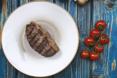 Kızarmış biftek, kiraz domatesleri, mavi ahşap masada tuz ve biber..