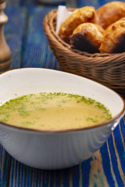 Beyaz kasede tavuk suyu çorbası ve mavi ahşap masadaki sepette pişmiş turtalar.