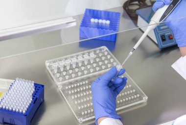Bir laboratuvar teknisyeninin ellerinde bir tüp kan örneği ve diğer numunelerle dolu bir raf var. Üzerinde çalışmak için kan tüpü örneği tutan laboratuvar teknisyeni. Corona virüsü salgını konsepti. Coronavirus aşısı gelişimi.