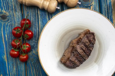 Kızarmış biftek, kiraz domatesleri, mavi ahşap masada tuz ve biber..