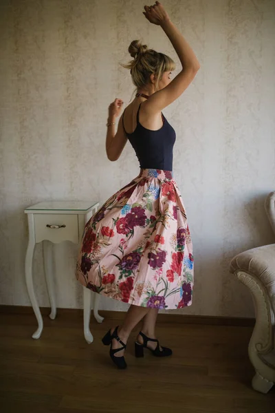 full length of stylish blonde woman in color floral skirt with red bow tie on neck posing indoors