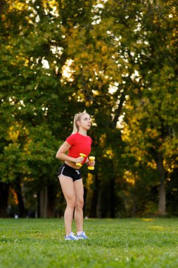 Genç bir kız spor için doğada dambılları olan bir yere gider. Spor salonundaki kadın parkta spor yapıyor..