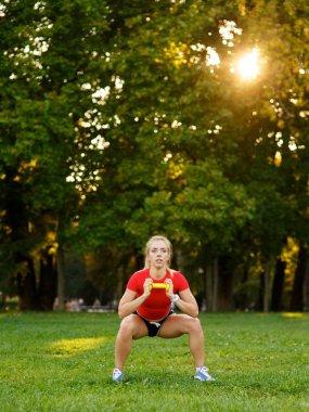 Genç bir kız spor için doğada dambılları olan bir yere gider. Spor salonundaki kadın parkta spor yapıyor..