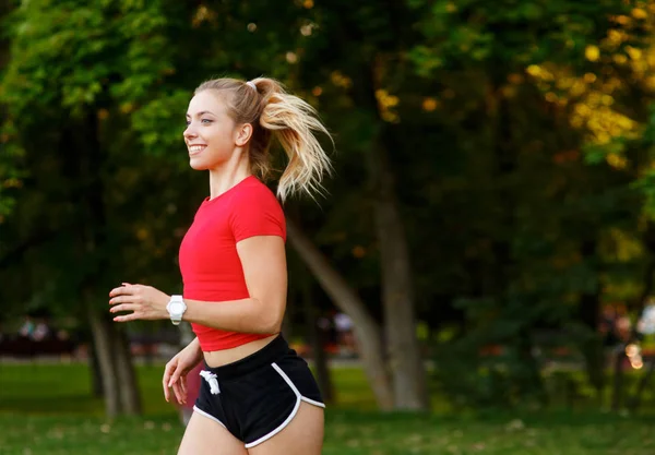 Młoda dziewczyna uprawia sport na świeżym powietrzu. kobieta w szortach i czerwony t-shirt biegnie w przyrodzie — Zdjęcie stockowe