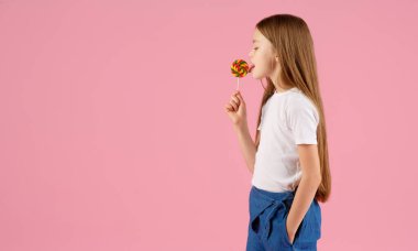 Kalp şeklinde lolipop tutan küçük bir kızın portresi pembe arka planda izole edilmiş kameraya bakıyor.