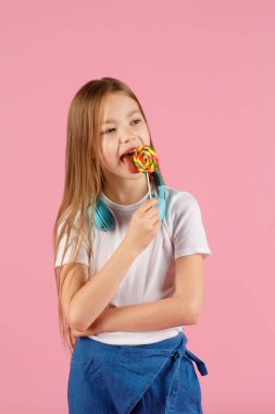Kalp şeklinde lolipop tutan küçük bir kızın portresi pembe arka planda izole edilmiş kameraya bakıyor.