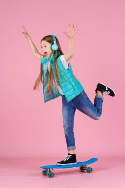 7-10 yaşlarında, ceketli güzel sarışın bir kız çocuğu. Pastel pembe arka plan çocuk portresi üzerinde izole bir şekilde poz veriyor. Çocukluk yaşam tarzı konsepti. Kopyalama alanı oluşturun. Mavi kaykayı tut