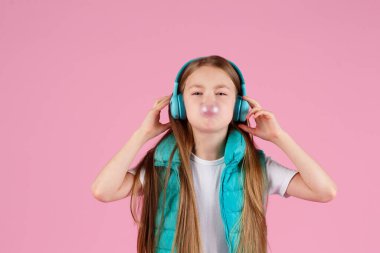 A little girl with headphones explodes pink chewing gum on a pink background. clipart