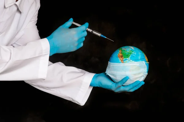 Globe earth in a medical mask and a syringe in the hands of the doctor close-up on a black background . The concept of the pandemic and the Coronavirus epidemic covid-19.