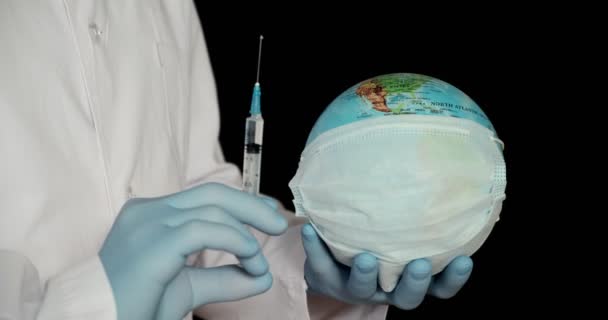 Globe jord i en medicinsk mask och en spruta i händerna på läkaren närbild på en svart bakgrund. Begreppet — Stockvideo