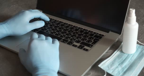 若いビジネスマンは、自宅のオフィスデスクに座っているラップトップコンピュータで働く医療用フェイスマスク手袋を着用しています。フリーランス — ストック動画