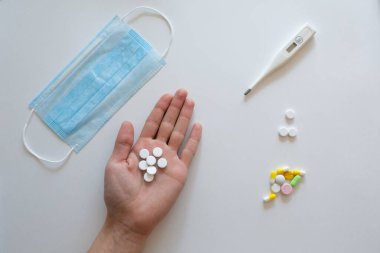 Doktorun ellerinde sıcaklığı ölçmek için elektronik bir termometre, koronavirüs ve tabletlerden koruyucu bir maske, mavi bir zemin üzerinde ilaçlar var. COVID-2019