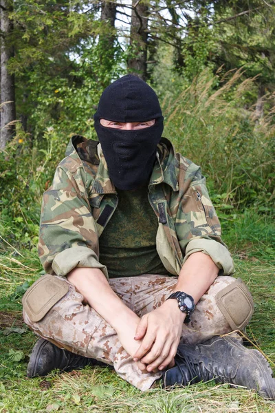 military man in camouflage and a black mask is sitting in the fo