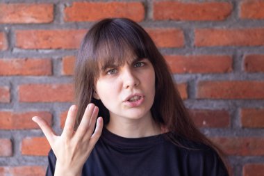 Annoyed brunette woman with bangs wearing a black t-shirt screaming due to conflict, showing all anger and anger on brick wall background