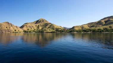 Coastline of mountains with green vegetation reflected  in blue ocean water at Labuan Bajo in Flores. clipart