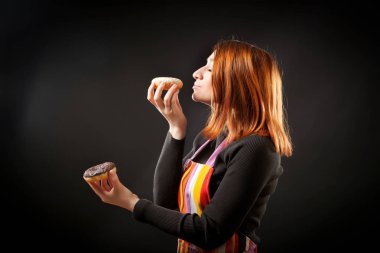 Kızıl saçlı kadın siyah boğazlı kazak ve mutfak önlük gülüyor ve zevk ile beyaz ve kahverengi siyah bir yalıtılmış zemin üzerine taze çörek burnunu çekmek