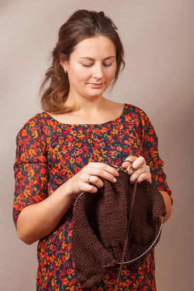 一个深色头发的孕妇在一张花印花的夏日礼服上的特写微笑着编织着编织的针从自然的雪利酒线在灰色的墙上的棕色毛衣 — 图库照片