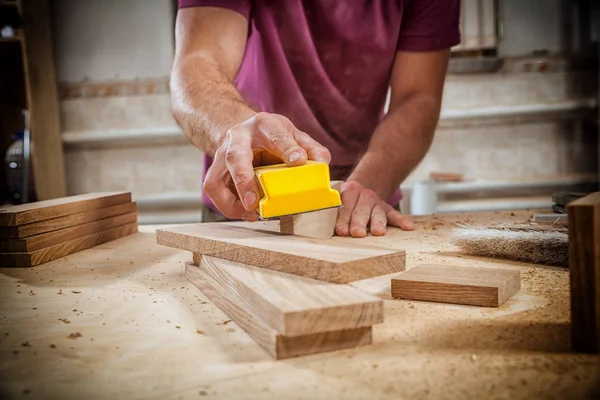Close-up of a young strong male carpenter builder in a purple working football polishes a wooden bar using sandpaper in the workshop, in the background a multitude of tools — Stock Image Close Young Strong Male Carpenter Builder Purple Working Football Polishes Royalty Free Stock Images