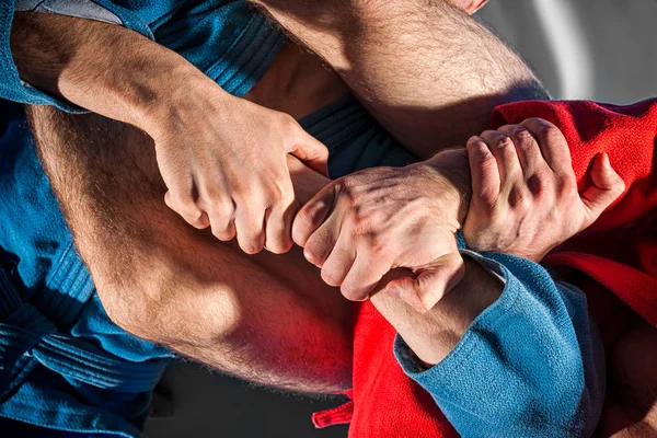 Close-up two wrestlers of sambo and jiu jitsu in a blue and red kimono doing . Man wrestler makes submission wrestling or armbar on gray tatami