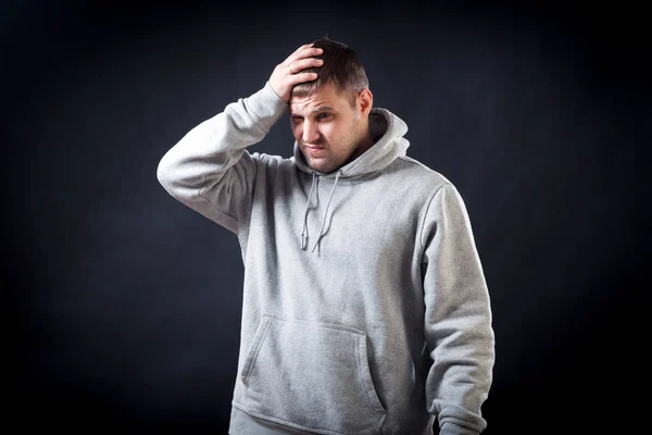 A young dark-haired man in a sporty gray sweatshirt was upset that he hit his head, now it hurts, his hands are holding on his head on a black isolated background