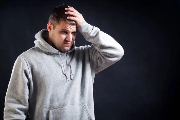 A young dark-haired man in a sporty gray sweatshirt was upset that he hit his head, now it hurts, his hands are holding on his head on a black isolated background