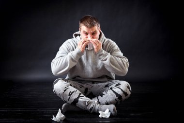 Üzgün delikanlı spor giysileri yere oturur ve o hasta, onun sümük akan üzgün, o bir peçete holding ve burnunu bir siyah izole arka planda üfleme
