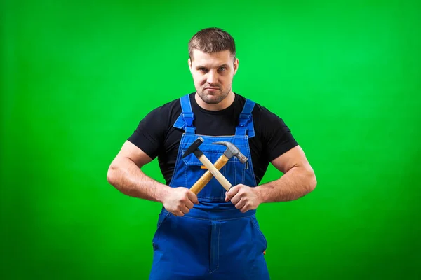 一个黑头发的男性建筑工人在一个黑色的 T恤和蓝色的建筑工作服正面摆正和持有两个木锤在一个绿色孤立的背景 — 图库照片