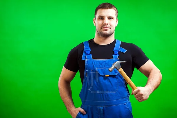 一个黑头发的男性建筑工人在一个黑色的 T恤和蓝色的建筑工作服漂亮的姿势和藏品在他的手木锤绿色隔离背景 — 图库照片