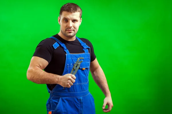 黑头发的男性建筑工人在黑色的 T恤和蓝色的建设整体持有一个新的灰色工具直管扳手在绿色隔离背景 — 图库照片