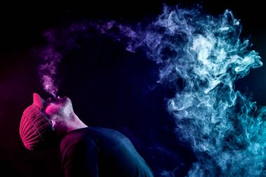 Genç bir adam bir gri ve siyah T-shirt maviden izole siyah bir arka plan üzerinde elektronik sigara duman kadar exhales