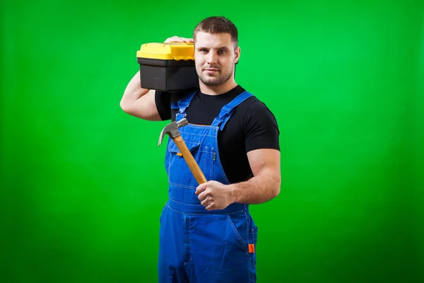 一个强壮的男子在黑色 T恤衫和蓝色建筑连身裤持有一个木制的锤子和一个盒子 他的肩膀上的建筑工具在绿色隔离背景 — 图库照片