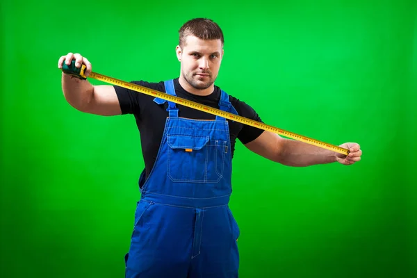 穿着黑色 T恤衫和蓝色建筑连身裤的男士木匠在绿色隔离的背景上使用长黄色的磁带规则 — 图库照片