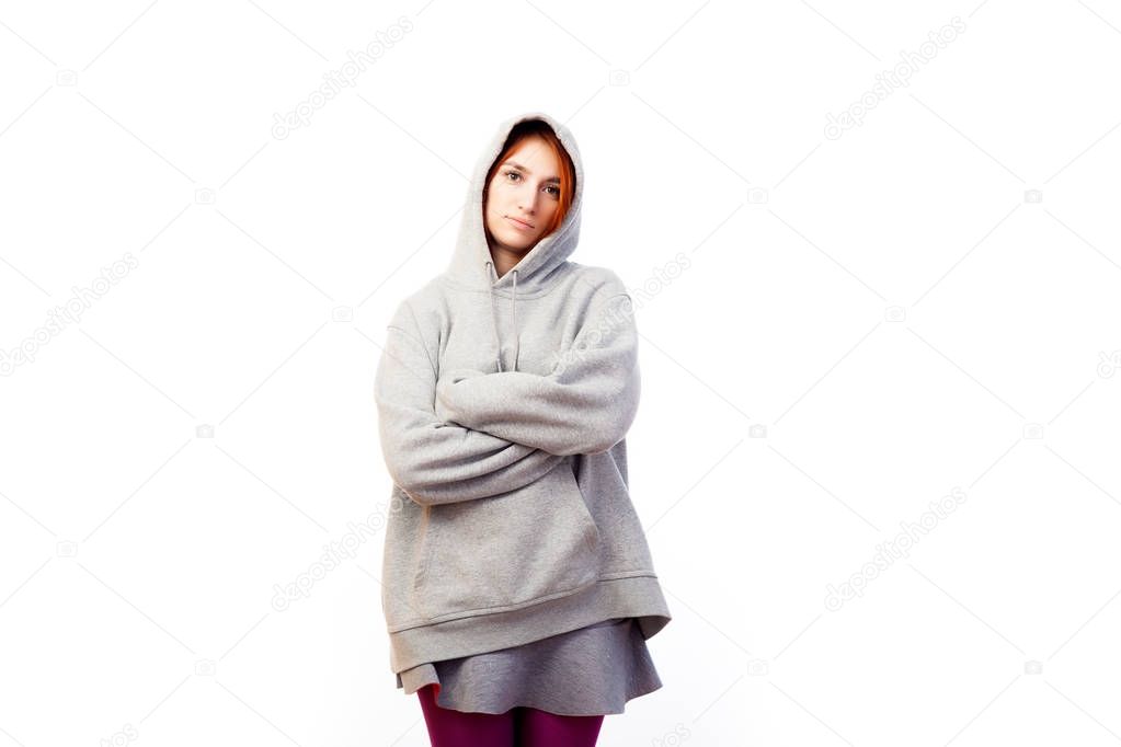 A young red-haired woman in a gray sweatshirt has put on a hood on her head, smiling and looking into the camera on a white isolated background