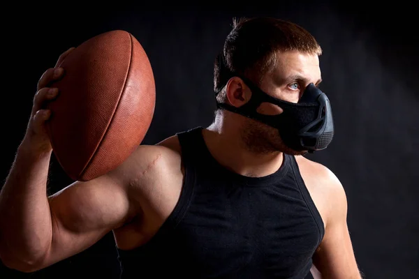 一个黑头发的男性运动员在一个黑色的训练面具 运动衫拿着橄榄球和挥动它在一个黑色孤立的背景 — 图库照片