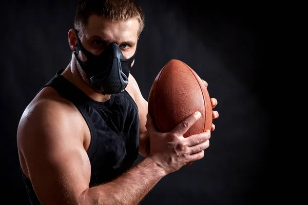 A dark-haired male athlete in a black training mask, a sports shirt holding a rugby ball on a black isolated background