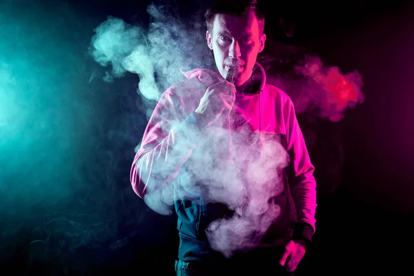 Man in sport hoody vaping an electronic cigarette.Isolated on black background.Around green and pink clouds of smoke