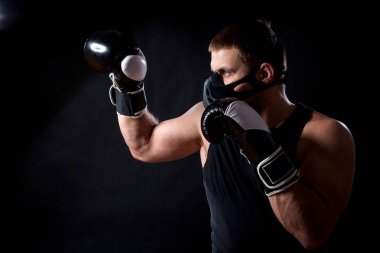 Sportif mavi gözlü genç adam spor siyah T-shirt, boks eldivenleri ve siyah eğitim maskesi giymiş bir siyah izole arka plan üzerinde boks