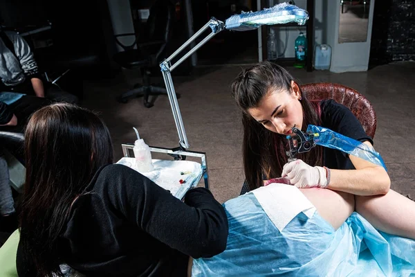 Dark haired woman tattoo master doing tattoo of a small bird of the tit on the hips with red tattoos on a young tattoo woman in a tattoo salon