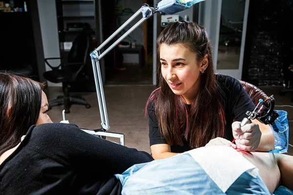 A young woman tattoo artist doing a tattoo with a tattoo machine under the bright light of a lamp on a beautiful woman with whom she talks in a dark workshop