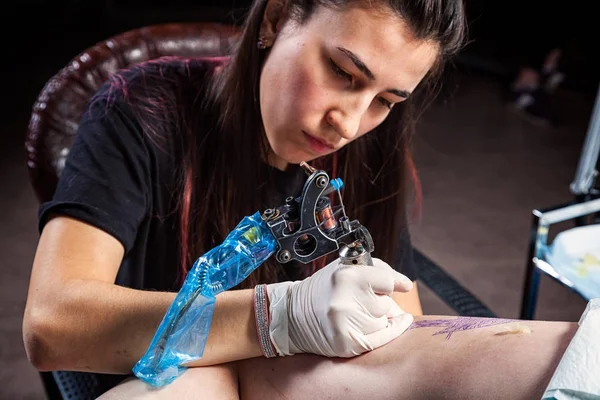 Young dark-haired woman tattoo master doing tattooed bird woman tattoo machine in dark workshop