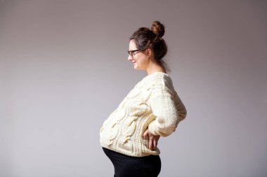 Güzel bir esmer genç kadın gözlük ve gülümseyen ve beyaz izole arka plan üzerinde poz örme süt renkli kazak ile geç gebelikte