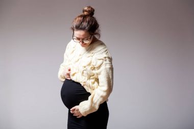 Geç gebelik ile gözlük ve örme süt renkli kazak poz ve midesini bir beyaz izole arka plan üzerinde tutan genç esmer güzel kadın
