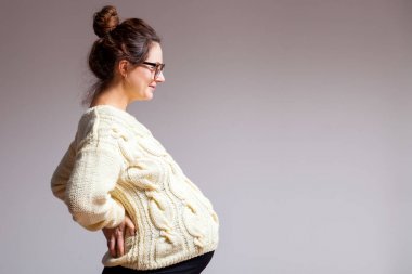 Güzel bir esmer genç kadın gözlük ve gülümseyen ve beyaz izole arka plan üzerinde poz örme süt renkli kazak ile geç gebelikte