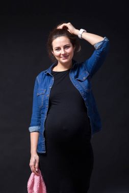 Young dark-haired beautiful woman on last pregnancy in jeans shirt and black dress smiles and holds in hands unbound pink sweater with knitting needles on a black isolated background clipart