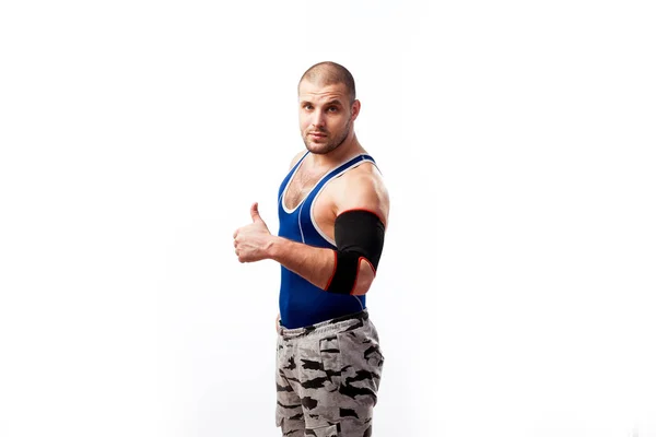 Young athletic male wrestler in blue shirt, sport pants and elbow pads stands and shows thumb up on white isolated background, side view