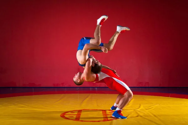 两名身穿蓝色和红色摔跤紧身衣的年轻人正在体育馆的一条黄色摔跤地毯上摔跤 — 图库照片
