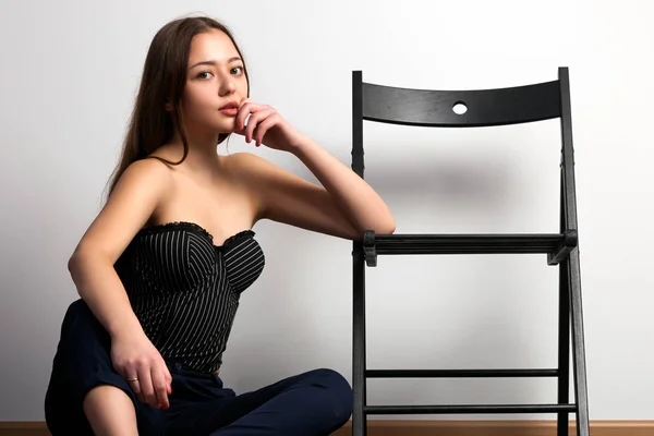 Asian young woman  in black top and classic black pants posing and sitting, black chair against a white wall background and black wooden floor
