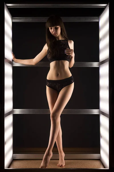 Young dark-haired woman in black  lingerie posing against white lights in black studio