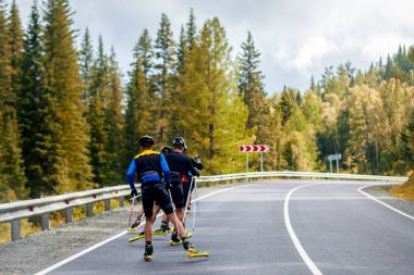 Takım biotlonists spor malzemeleri içinde Koruyucu baretler eller Kayak Polonyalılar tutmak silindir kayaklar, Altay dağlık alanda bir yaz gününde orman boyunca asfalt yol boyunca kayak gitmek