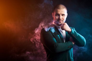 Genç yakışıklı adam baş beyaz gömlek ve gülümseyen düşünceli kamera karşısında kırmızı ve mavi duman mendil siyah bir izole arka planda bakarak ve yeşil ceket